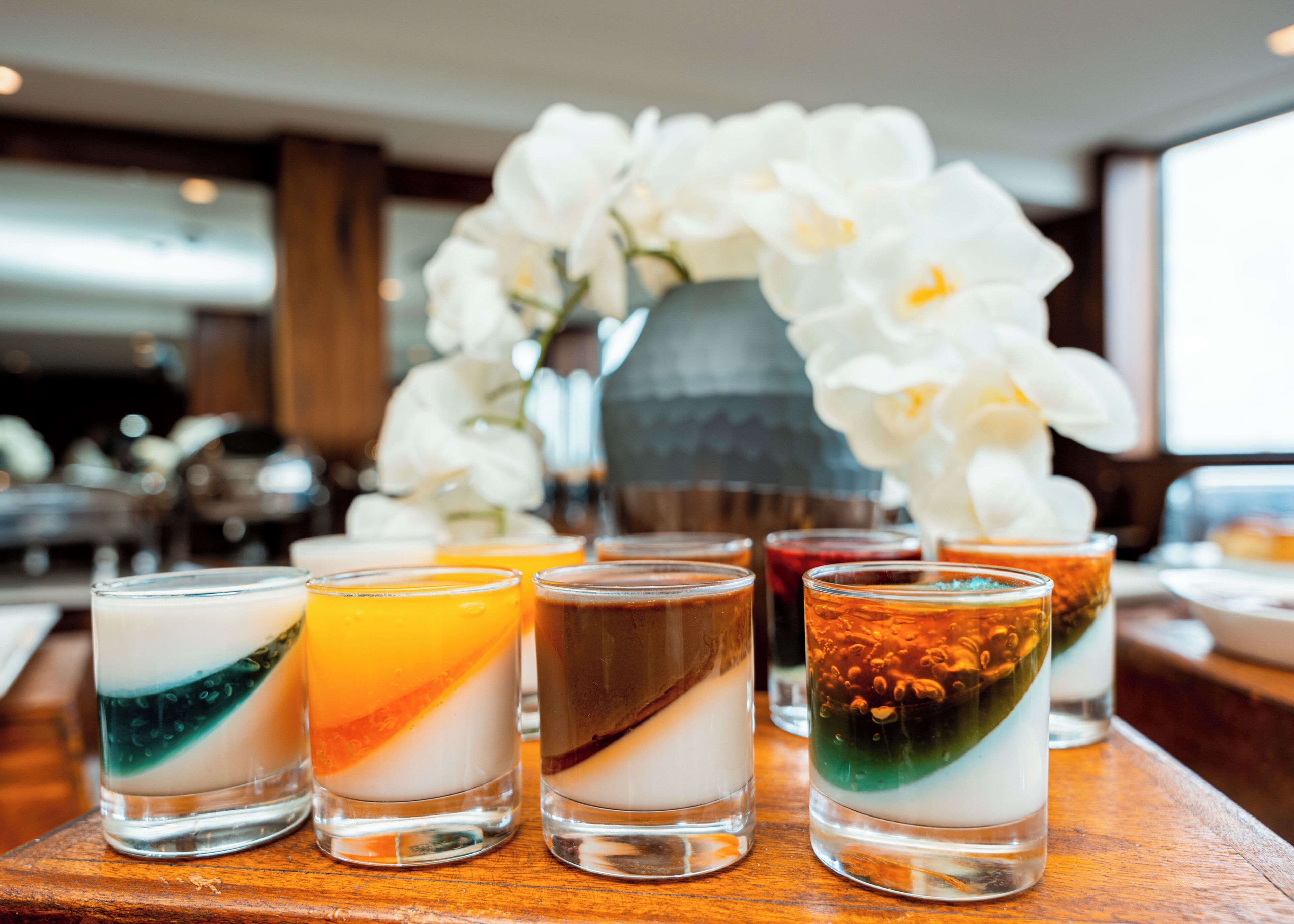 Una zona de bar con varios vasos de bebidas en capas y un jarrón con flores blancas al fondo.