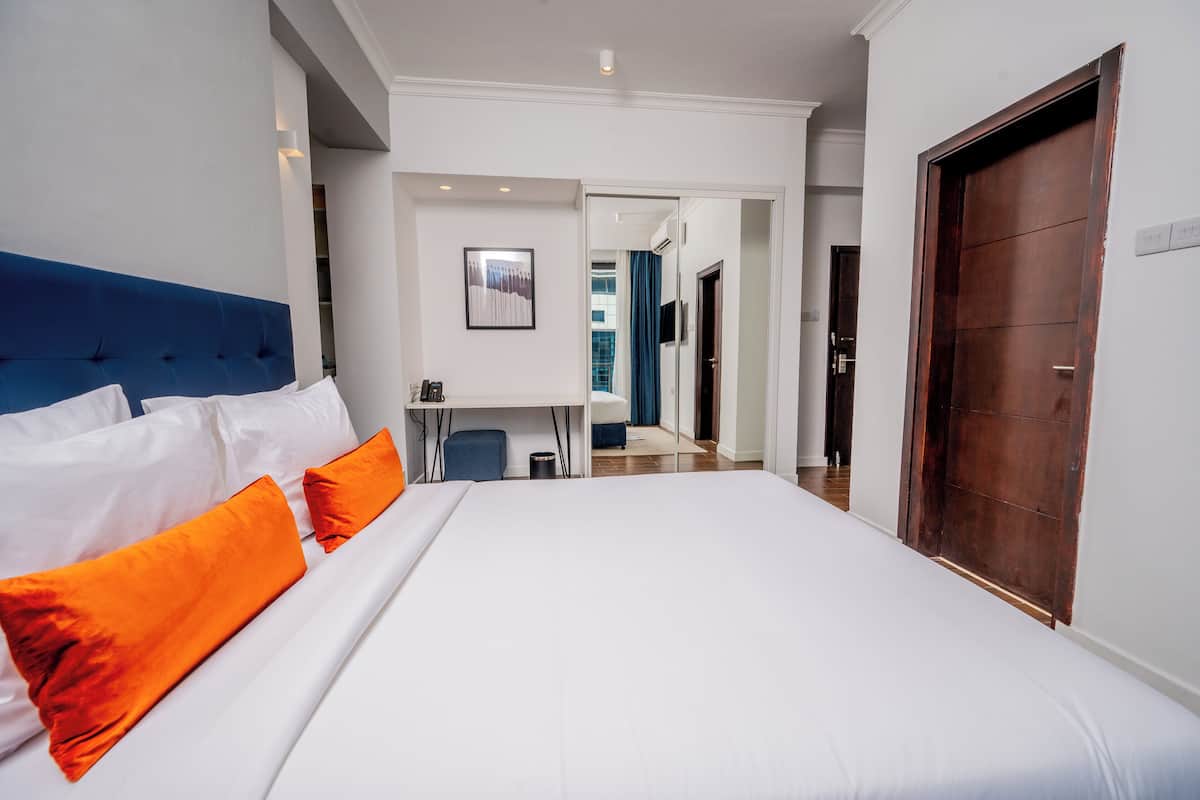 Ett hotellrum med en stor säng, vita och orange kuddar, ett skrivbord med en stol och en dörr av trä.