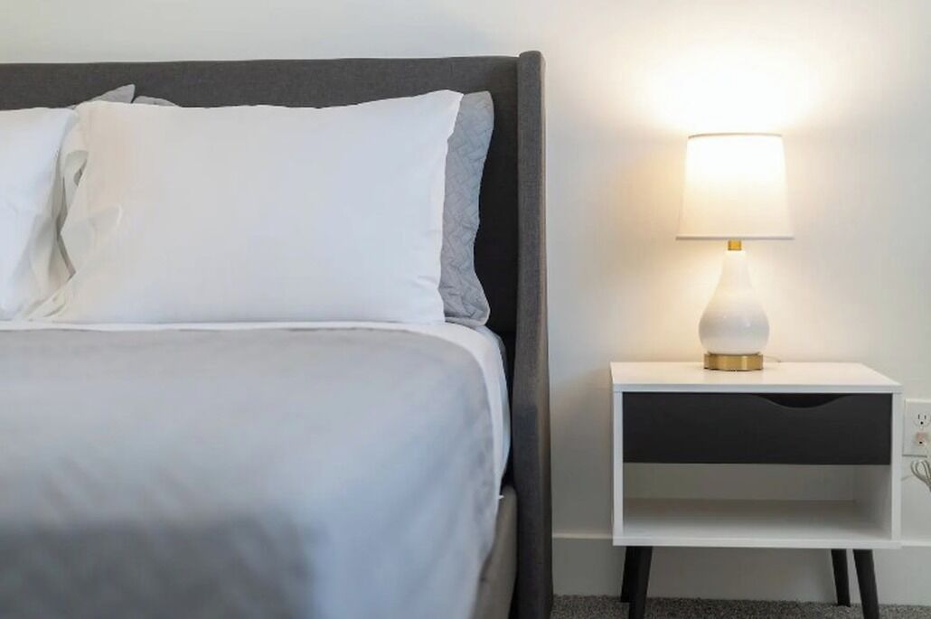 A neatly made bed with white bedding and a grey headboard, a bedside table with a lamp, and a power outlet.