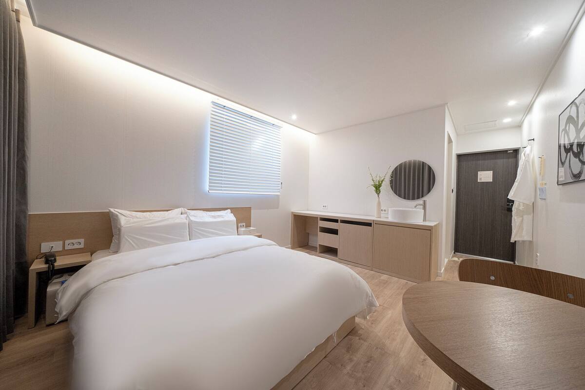 A modern hotel room with a large bed, bedside tables, a desk, a vanity with a mirror, and a door.