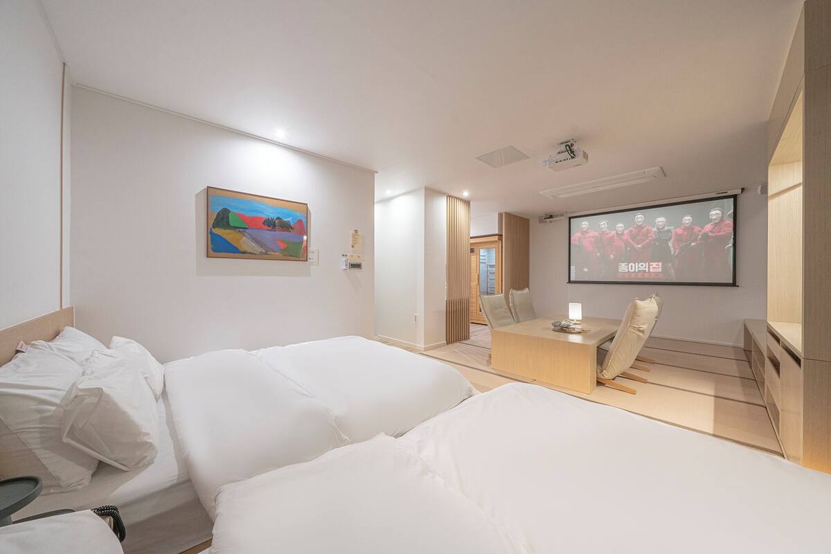 A modern hotel room with a large bed, a desk, and a TV screen displaying a group of people.