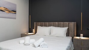 Una cama bien tendida con sábanas blancas y toallas enrolladas, un cabecero oscuro y mesitas de noche con lámparas.