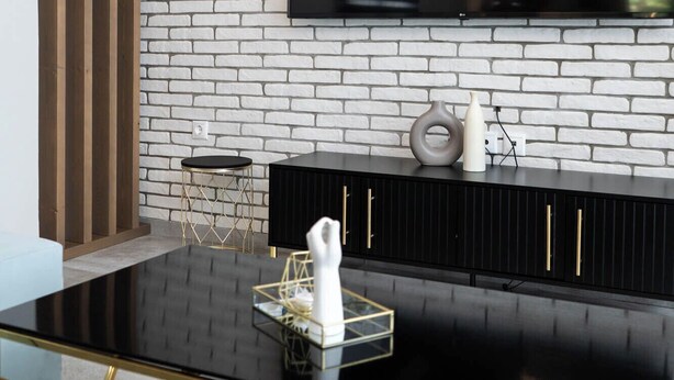 Una sala de estar moderna con una mesa de centro de vidrio negro, un televisor negro sobre una pared de ladrillo blanco y un mueble negro con tiradores dorados.