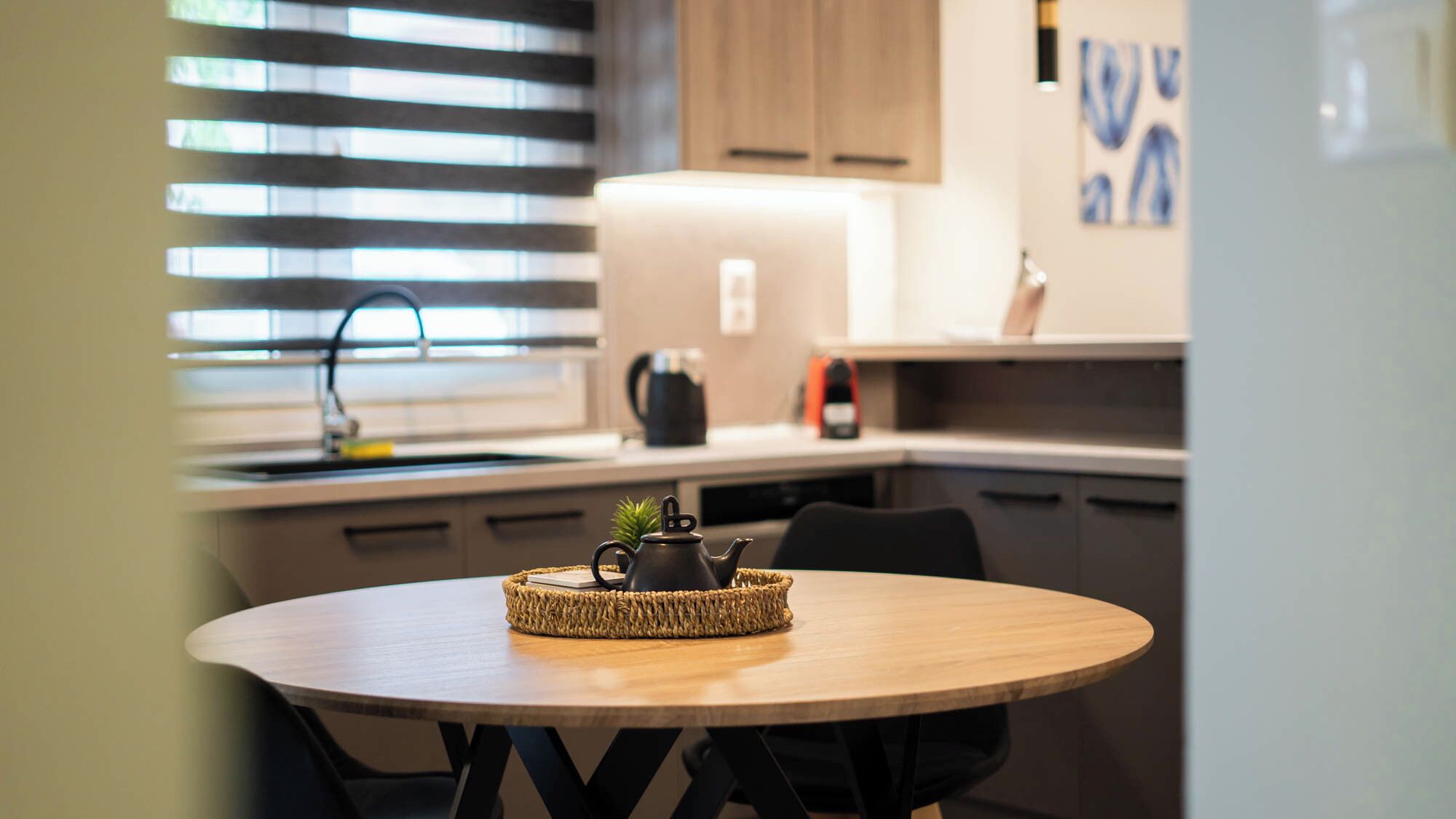 A modern kitchen with a round wooden table, a woven placemat, a teapot, and a small plant.
