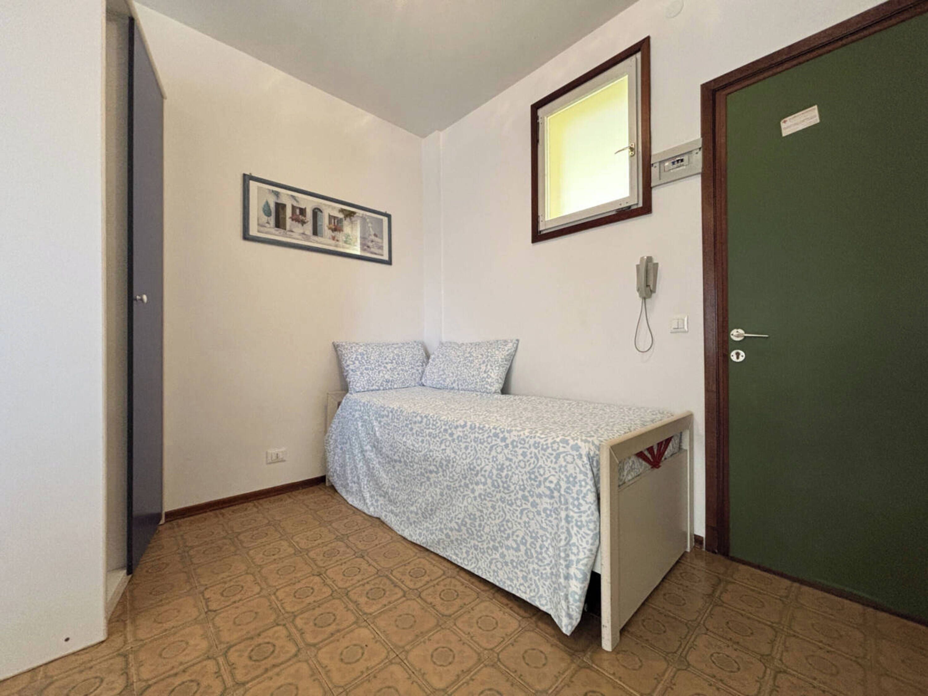 A small, single-bed room with a patterned bedspread, a framed picture on the wall, a telephone, and a green door.