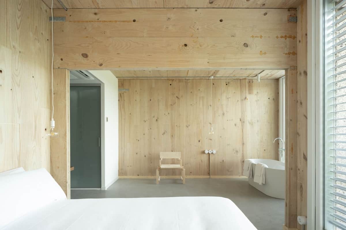 A minimalist bedroom with a wooden ceiling, a single bed, a small wooden chair, a built-in sink, and a window with blinds.