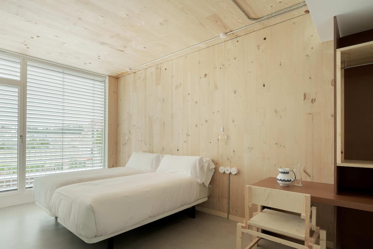A minimalist bedroom with a wooden wall, a bed, a small wooden table, and a window with blinds.