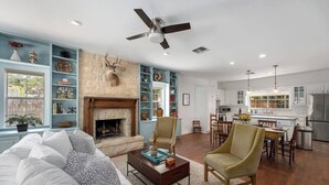 A spacious living room with a fireplace, a dining area, and a kitchen with white cabinets and a stainless steel refrigerator. - Revins Haus - Fire Pit & Walk to Main St (Fredericksburg)