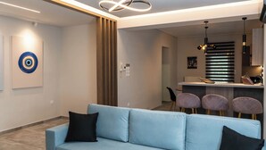 A modern living room with a light blue sofa, a decorative wall piece, and a minimalist kitchen area with a bar. - Mythic Meteora (ΚΑΛΑΜΠΑΚΑ)