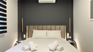 A modern hotel room with a large bed, two bedside tables, a wall-mounted air conditioner, and a small round table with a phone and remote control. - Mythic Meteora (ΚΑΛΑΜΠΑΚΑ)