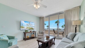 Un salón amplio con un gran ventanal, ventilador de techo, televisor de pantalla plana y balcón con vista a palmeras.