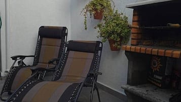 Een terras met twee loungestoelen, een kleine bakstenen barbecue en potplanten.