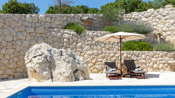 A swimming pool with stone walls and a large rock formation. There are two lounge chairs with an umbrella. - Private villa Iko with swimming pool and view on mountain Biokovo (Zadvarje)