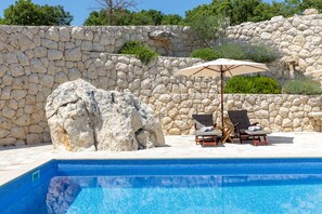 Una piscina con pareti in pietra e una grande formazione rocciosa. Sono presenti due lettini da spiaggia con un ombrellone.