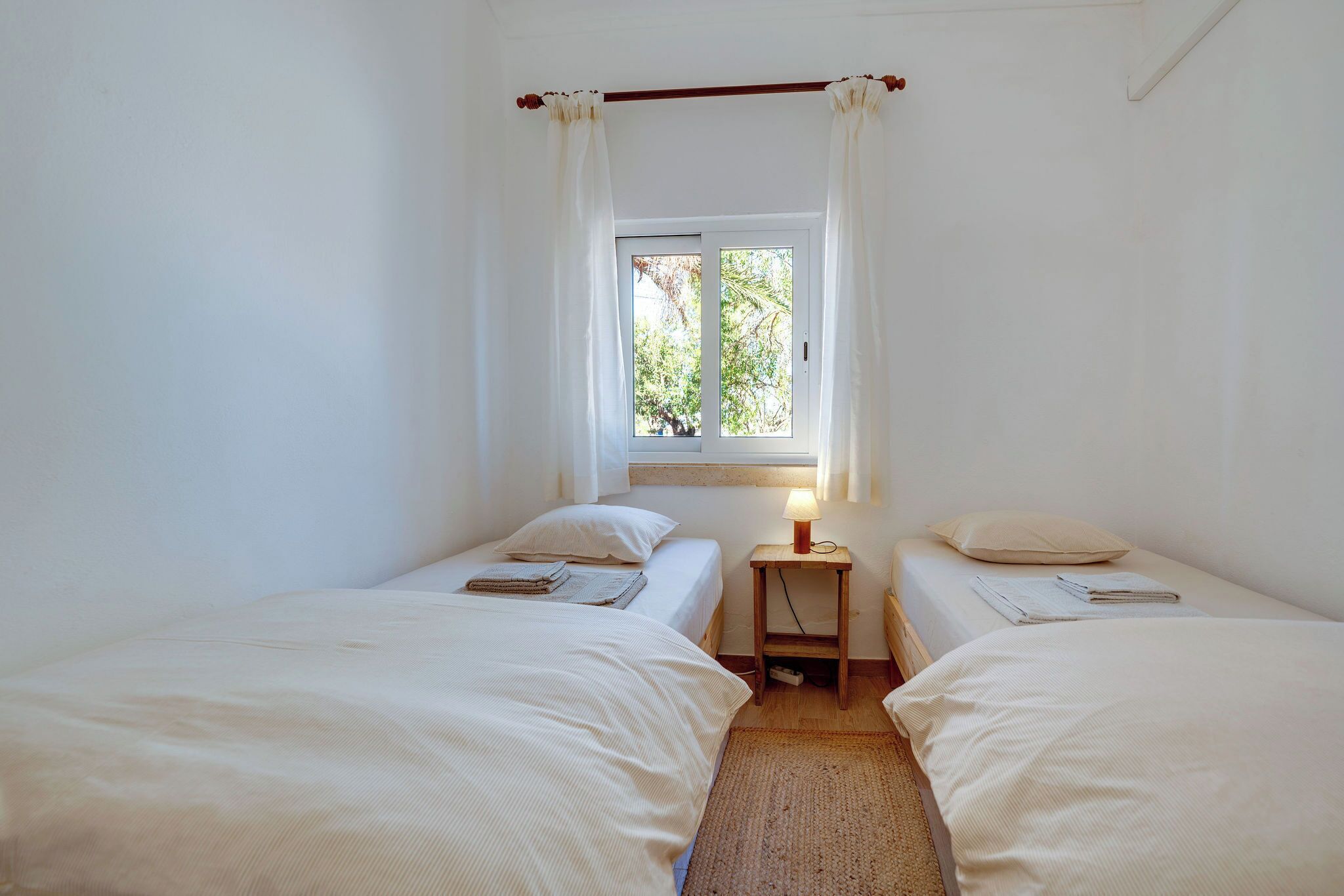A small, clean room with two single beds, a window with curtains, and a bedside table with a lamp.