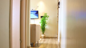 Ein Flur mit Holzboden, einer Topfpflanze, einem beigefarbenen Sofa und einem an der Wand montierten Fernseher.