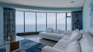 A modern living room with a large white sofa, a glass coffee table, and a view of the ocean.