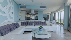 A modern living room with a large sectional sofa, a glass coffee table, and a view of the kitchen and outdoor balcony.