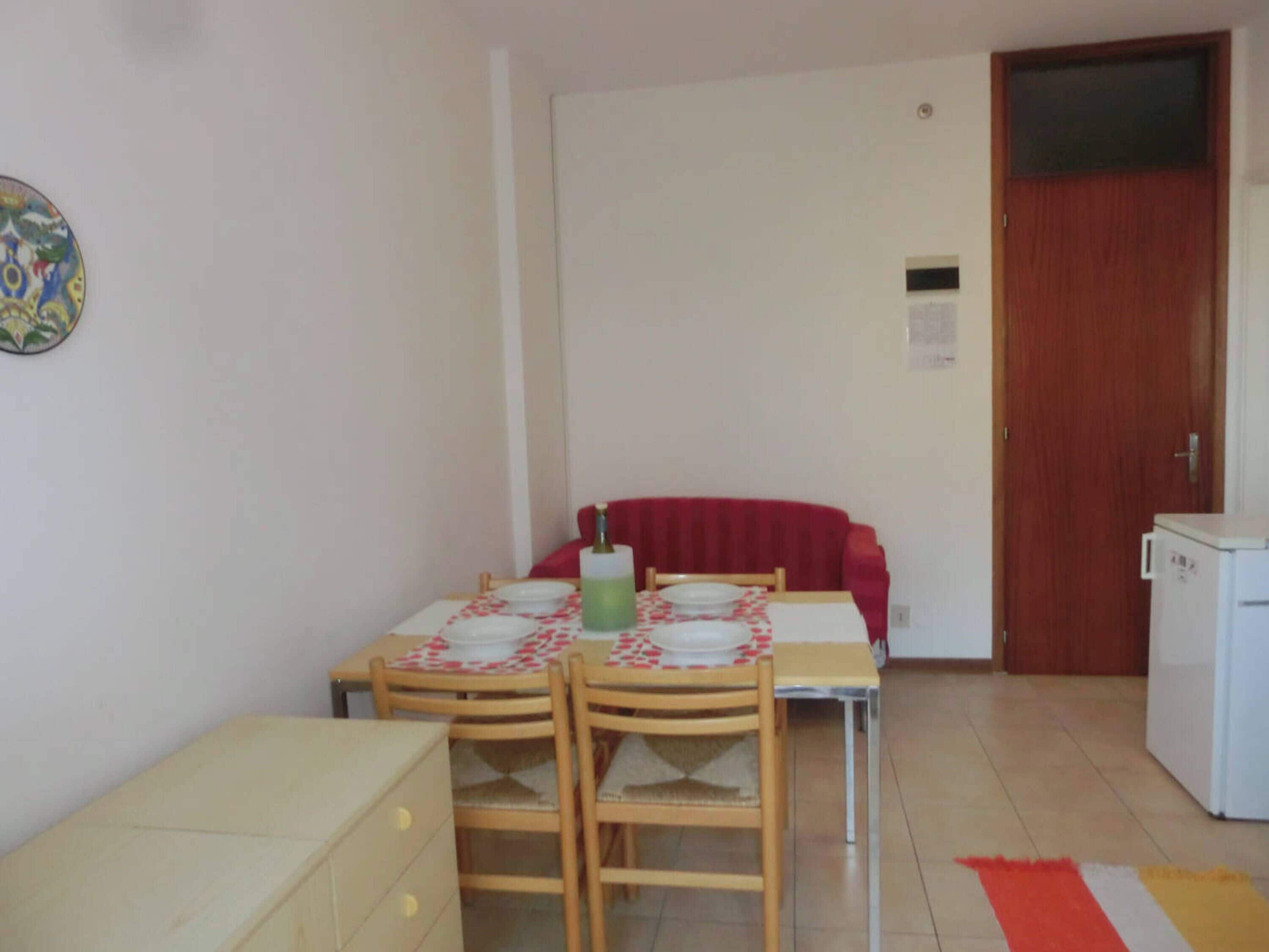 A small, compact room with a dining table set for two, a red sofa, a small cabinet, and a refrigerator.