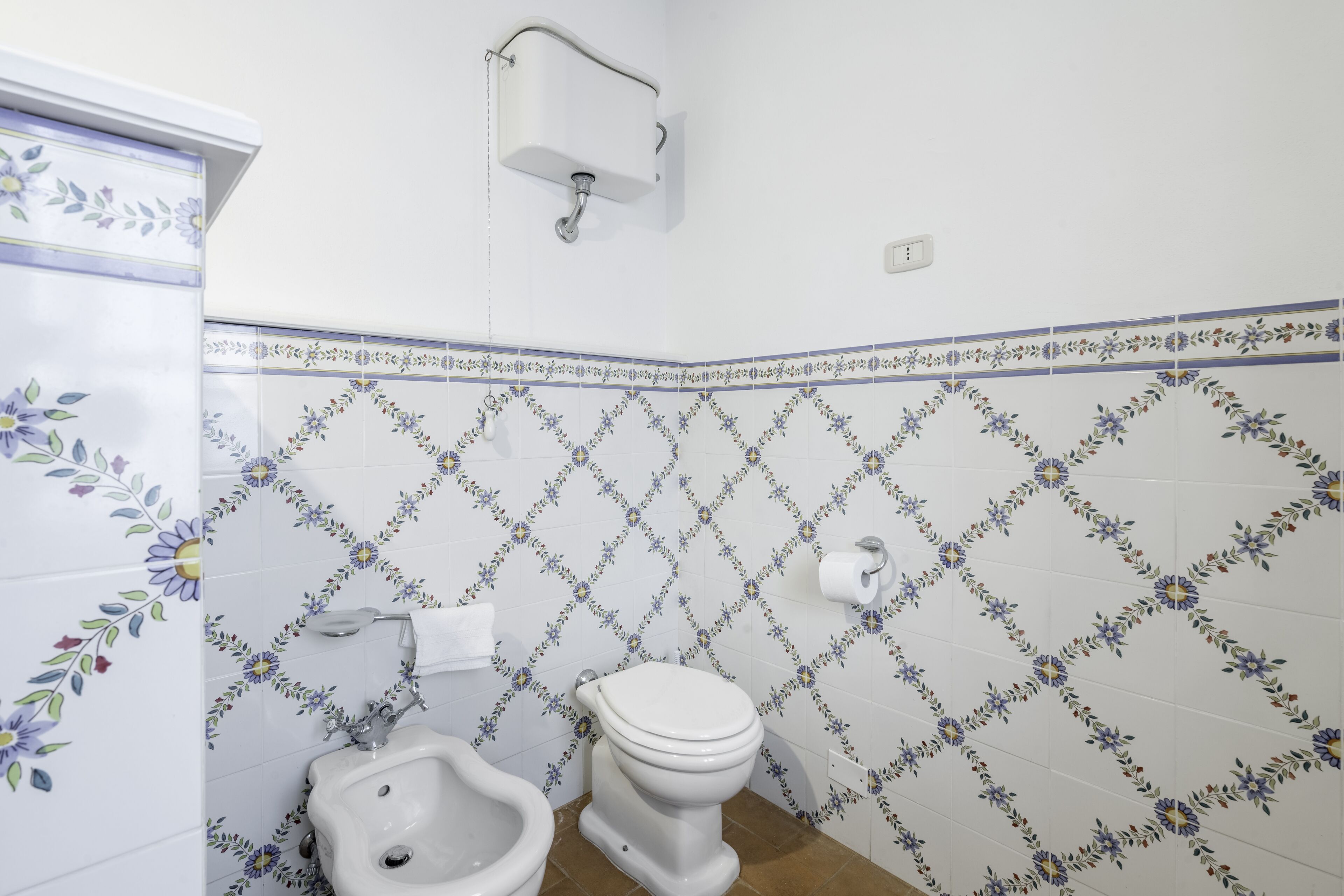 A bathroom with a toilet, bidet, and sink, featuring floral patterned tiles and a white wall.