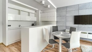 A modern kitchen with white cabinetry, a central island with seating, and a flat-screen TV mounted on the wall. - MARGI Eclipse Apartament (Szczecin)