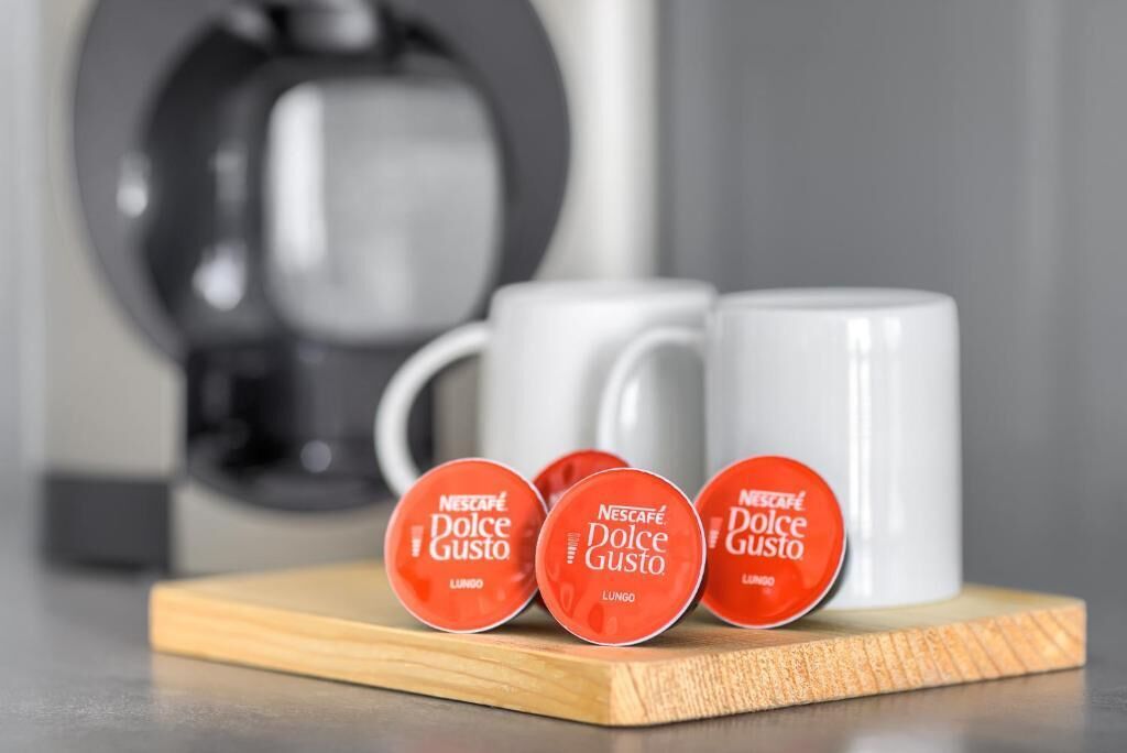Drei Nescafe Dolce Gusto Kapseln auf einem Holzuntersetzer, eine weiße Tasse und eine Kaffeemaschine im Hintergrund.