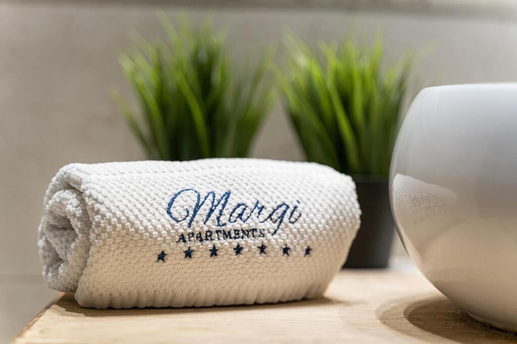 A folded towel with "Margi Apartments" embroidered on it, placed on a wooden surface next to a bowl.
