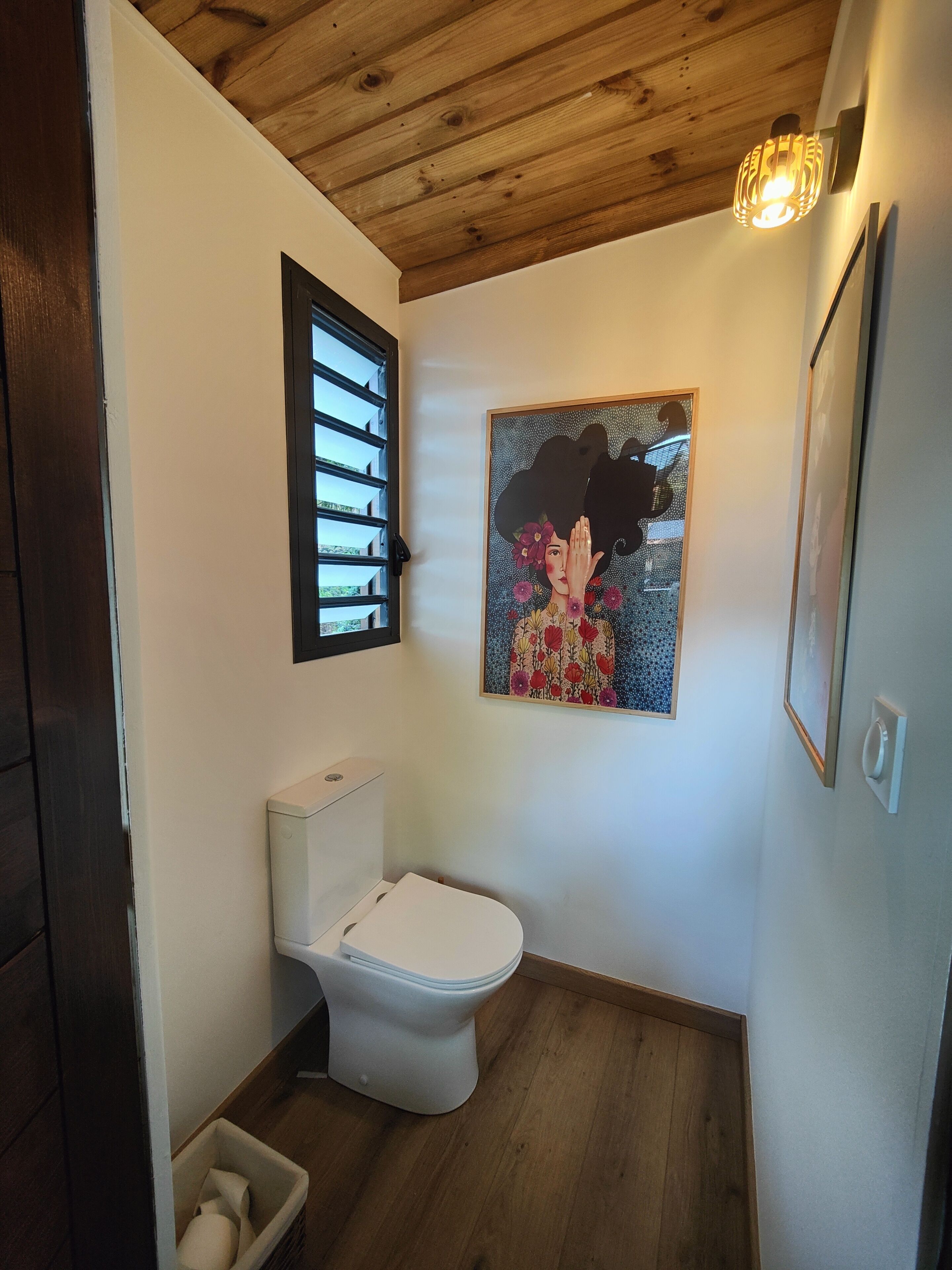 Een kleine badkamer met een wit toilet, een houten vloer, een plafond met zichtbare balken, een wandlamp en een lijstwerk afbeelding boven het toilet.