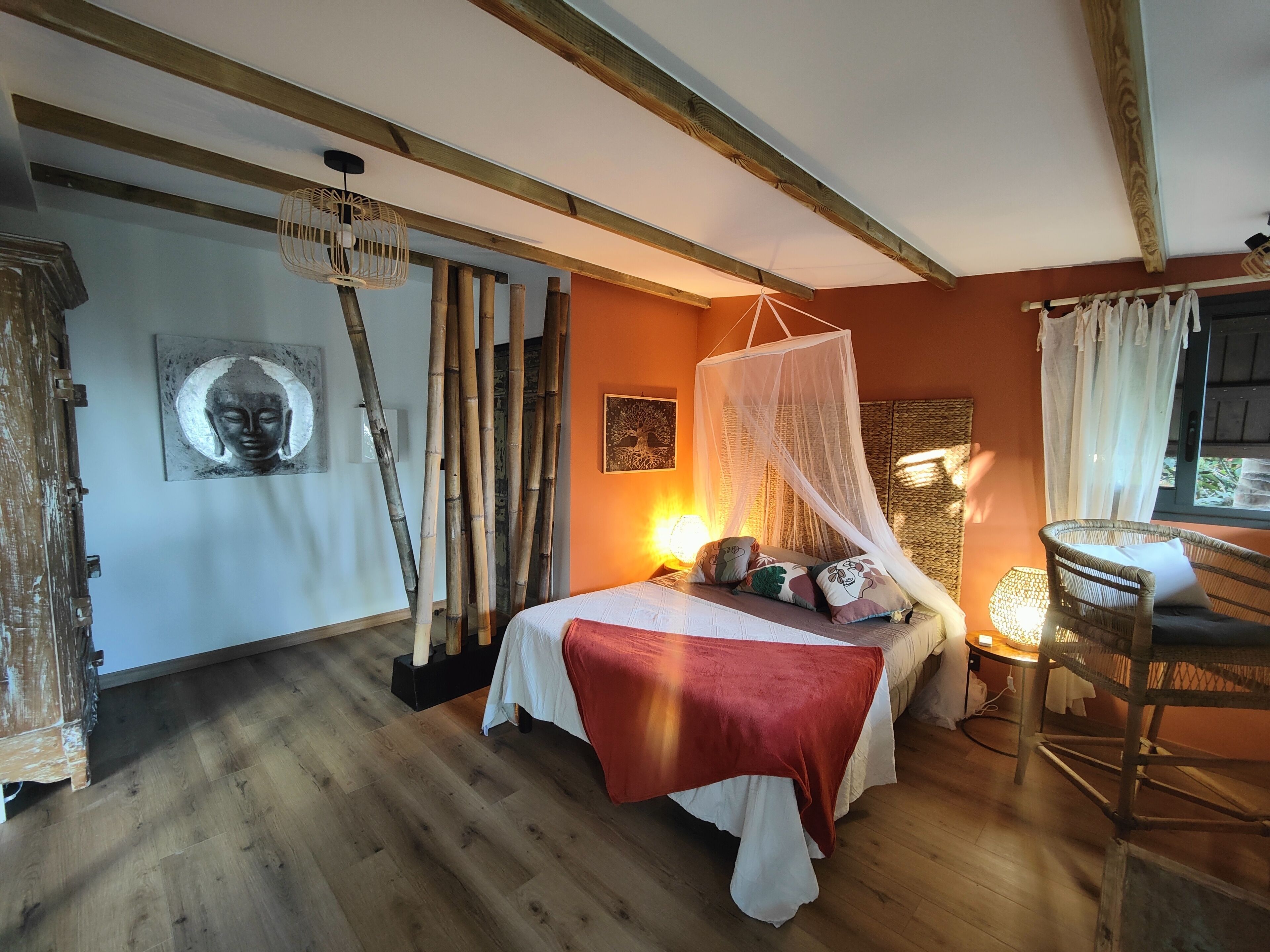 Ein Schlafzimmer mit einem Bett, einem Nachttisch, einem Sessel, einem Wandventilator und einem Fenster mit Vorhängen.