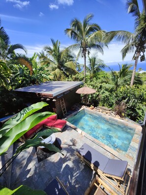 Un espace piscine doté de chaises longues et d’une terrasse en bois, entouré d’une végétation tropicale et offrant une vue dégagée sur l’océan.