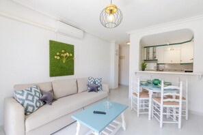 Un salon comprenant un canapé, une table basse, une table à manger avec des chaises, un coin cuisine et un luminaire suspendu.