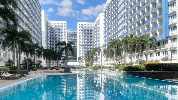 A swimming pool surrounded by high-rise buildings with balconies, palm trees, and a clear sky. - 2BR Poolview  for 6-10 pax at Sea Residences Mall of Asia 402403 (Pasay)