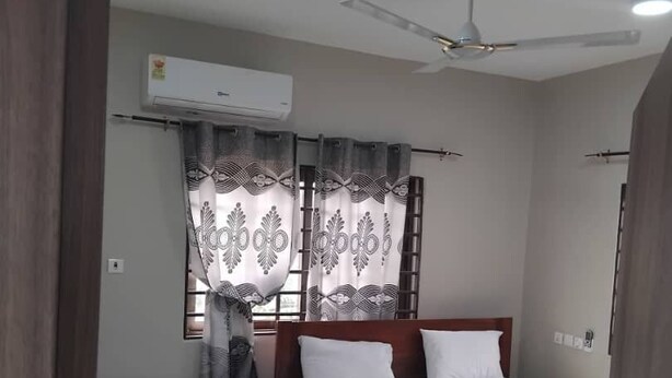 Una camera da letto con un letto, un comodino, una sedia, una finestra con tende, un condizionatore d'aria e una ventola a soffitto.