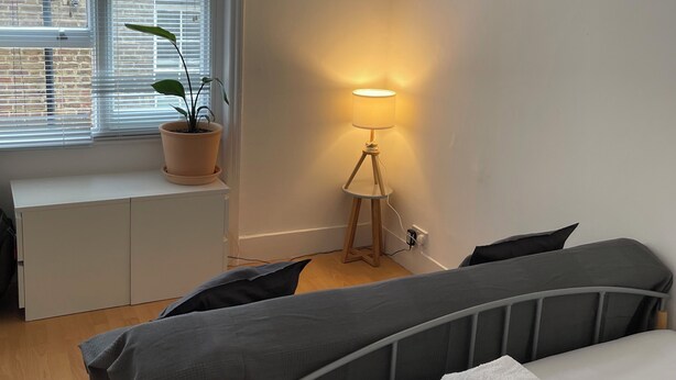 A bedroom with a bed, bedside tables, a lamp, a window with blinds, and a potted plant. - SOOTHEUS - SOHO7 (London)