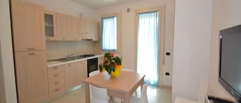 A modern kitchen with light wooden cabinets, a dining table with chairs, and a TV on the right.