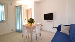 A modern living room with a blue sofa, a dining table with chairs, a TV mounted on the wall, and a door with curtains. - Modern Flat in Central Location (Rosolina)