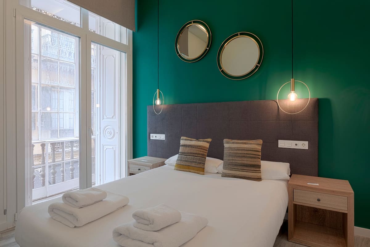 A hotel room with a large bed, two bedside tables, a window with a view of buildings, and two wall-mounted mirrors.
