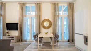 A modern living room with a dining table, chairs, a flat-screen TV, and large windows with curtains. - Fresh Velazquez Apartments (Málaga)