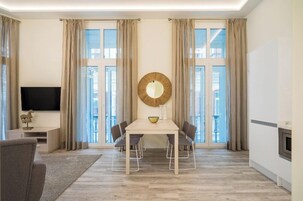 A modern living room with a dining table, chairs, a flat-screen TV, and large windows with curtains.