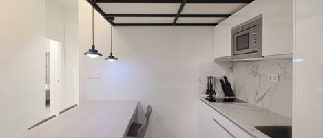 Una cucina compatta con un piano di lavoro bianco, un forno a microonde integrato e un lavandino. Si nota un tavolo da pranzo con sedie e illuminazione a sospensione.