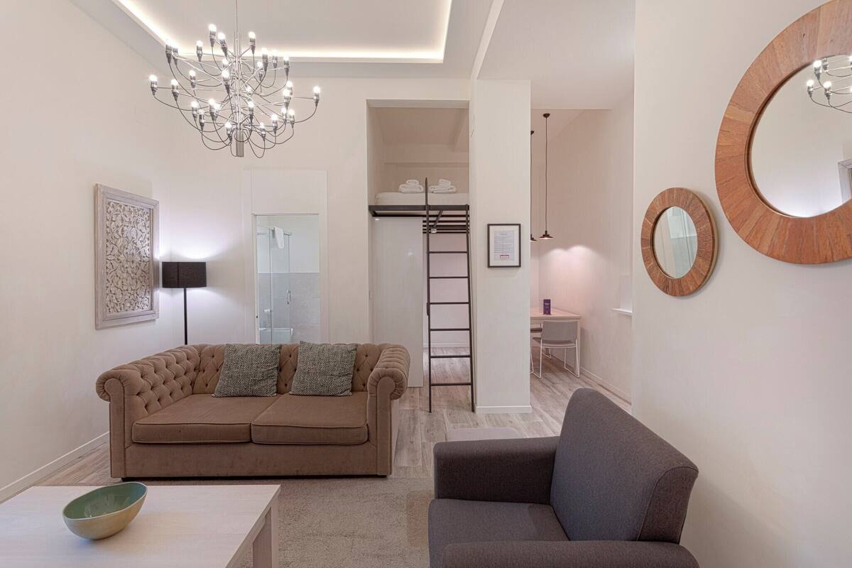 A modern living room with a brown sofa, a grey armchair, a chandelier, and a large mirror.