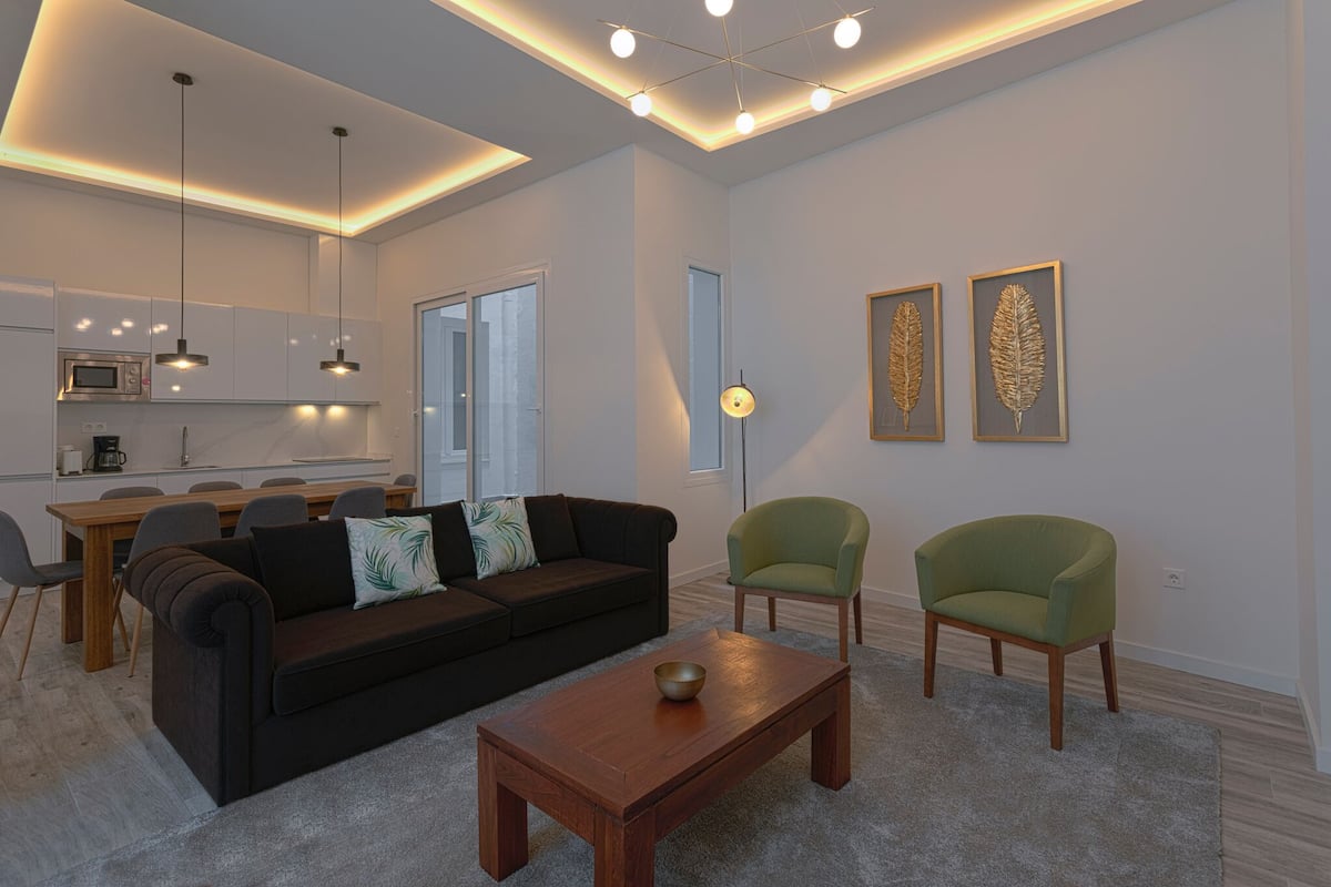 A modern living room with a dark sofa, two green armchairs, a wooden coffee table, and a dining area with a wooden table and chairs.