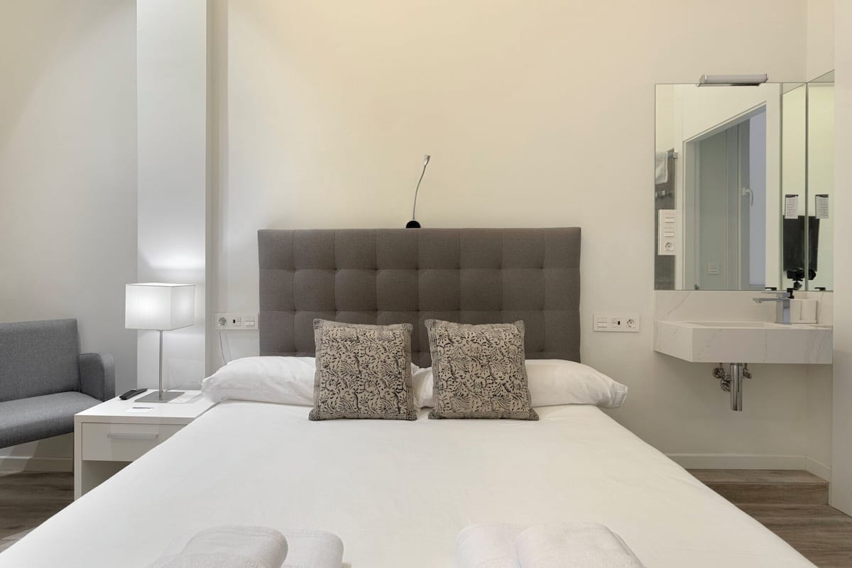 A modern hotel room with a large bed, a bedside table, a lamp, a chair, and a bathroom with a mirror and sink.