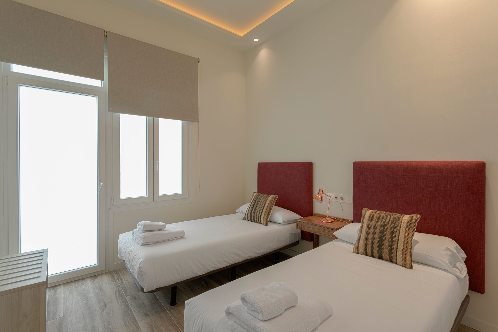 A hotel room with two beds, a small table with a lamp, and a window with a view.