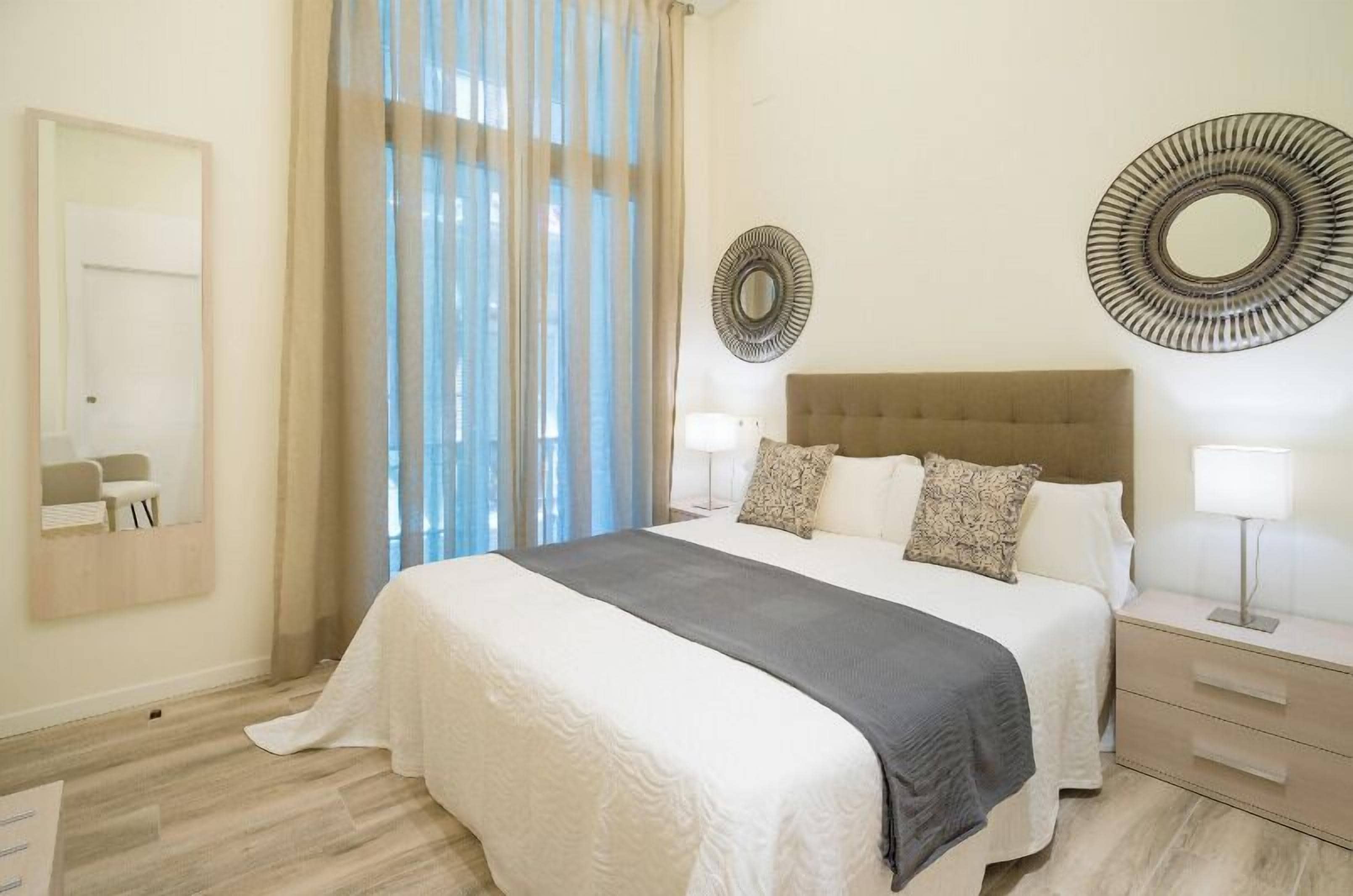 A bedroom with a large bed, a bedside table with a lamp, a mirror on the wall, and sheer curtains.