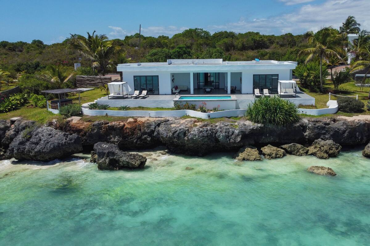 A modern house with a pool, surrounded by tropical vegetation and a rocky shoreline.