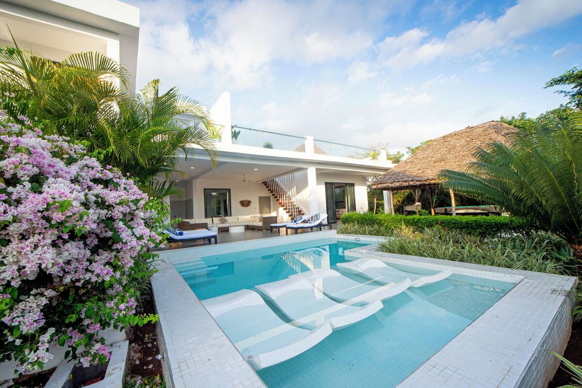 A modern pool area with a clear blue pool, white lounge chairs, and a staircase leading to a covered upper level.