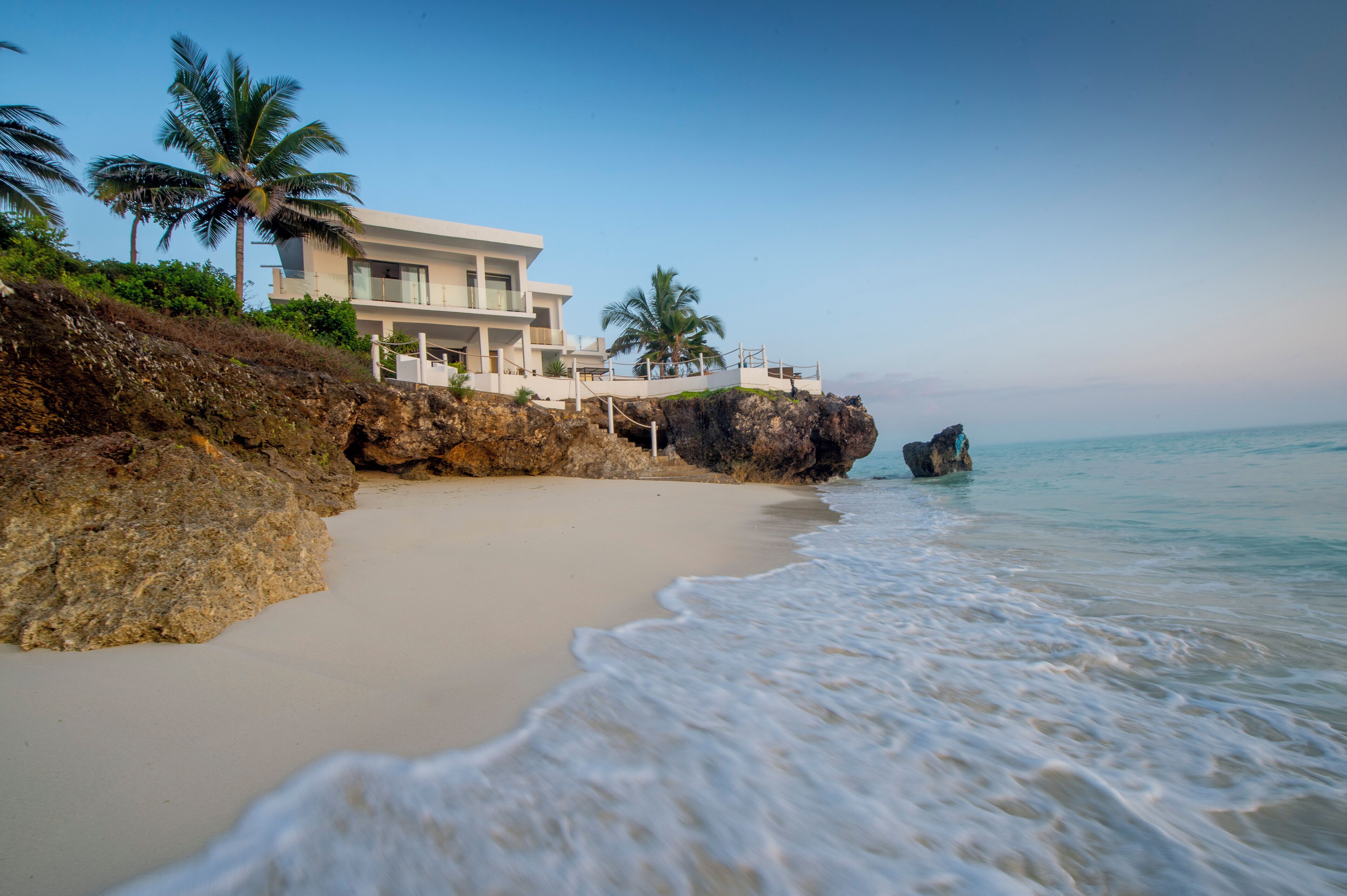 A beachfront property with a modern house, palm trees, and a rocky shoreline.