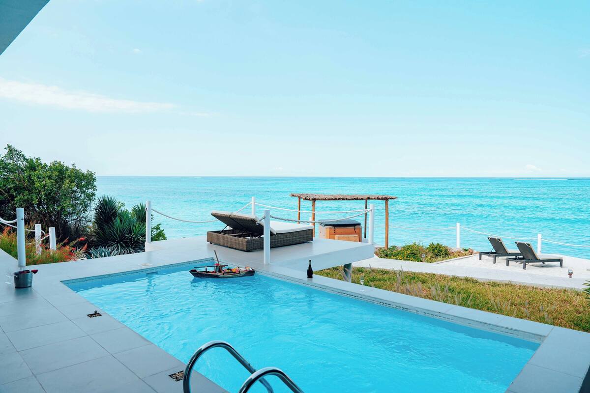 A pool with a view of the ocean, lounge chairs, and a small boat.