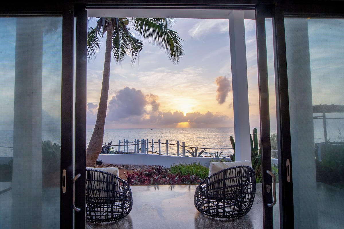 A view through a glass door of a balcony with two wicker chairs, a palm tree, and a sunset over the sea.